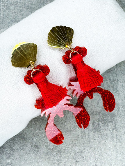 Dieser Ohrring ist gefertigt aus einem Edelstahl-Ohrsstecker in Form einer Venus-Muschel, einem rot glitzerndem Acryl-Lobster-Anhänger und einer roten Quaste.