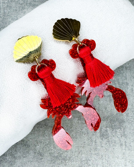 Dieser Ohrring ist gefertigt aus einem Edelstahl-Ohrsstecker in Form einer Venus-Muschel, einem rot glitzerndem Acryl-Lobster-Anhänger und einer roten Quaste.