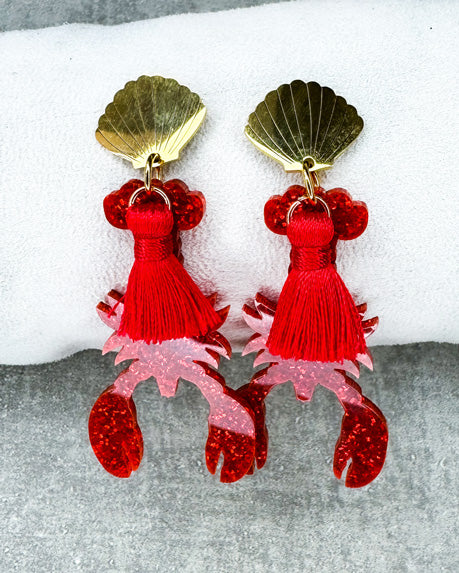Dieser Ohrring ist gefertigt aus einem Edelstahl-Ohrsstecker in Form einer Venus-Muschel, einem rot glitzerndem Acryl-Lobster-Anhänger und einer roten Quaste.
