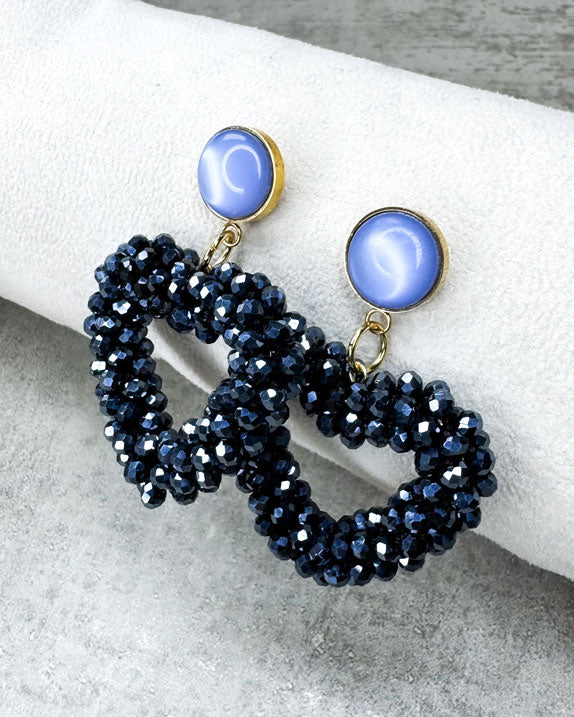 Dieser Ohrring ist gefertigt aus einem Cabochon-Stecker mit einem hellblau, glänzenden Stein und einem glitzernden Facetten-Herz-Anhänger in der Farbe "Silver Black.