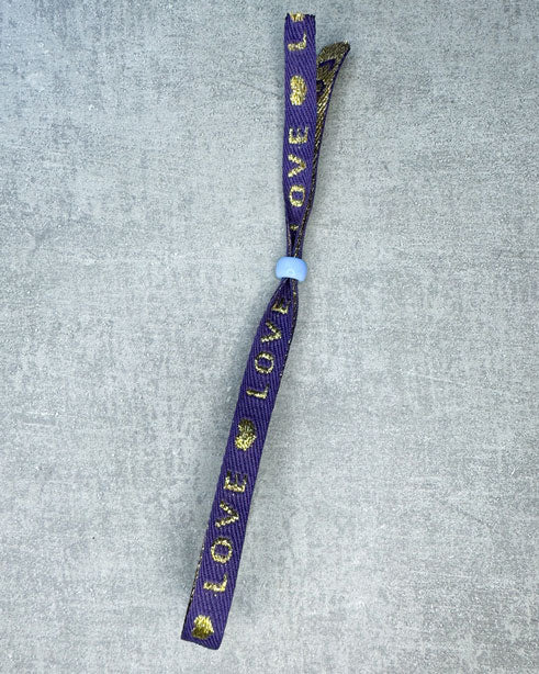 Dieses-Armband besteht aus sehr haltbarem Polyester und hat die Aufschrift "PURPLE LOVE". Die goldfarbene Stopper-Perle ist aus rostfreiem Stahl.