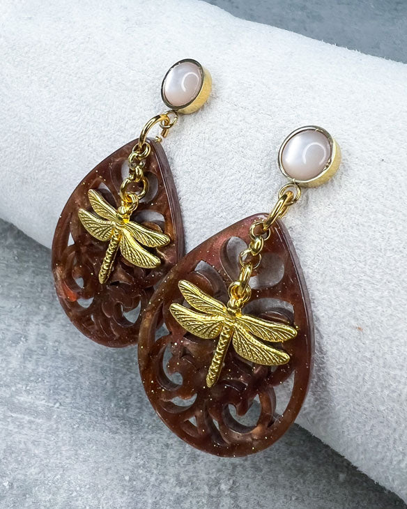 Diese Ohrringe sind individuell gestaltet und handgefertigt von uns!  Diese Ohrringe sind gefertigt aus: einem goldfarbenem Cabochon Stecker mit einem babyrosafarbenem Stein, einem Ornament-Anhänger in Braun-Glitzer und einem goldfarbenen Libellen-Anhänger.