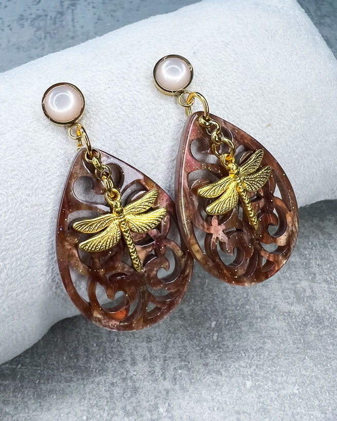 Diese Ohrringe sind individuell gestaltet und handgefertigt von uns!  Diese Ohrringe sind gefertigt aus: einem goldfarbenem Cabochon Stecker mit einem babyrosafarbenem Stein, einem Ornament-Anhänger in Braun-Glitzer und einem goldfarbenen Libellen-Anhänger.