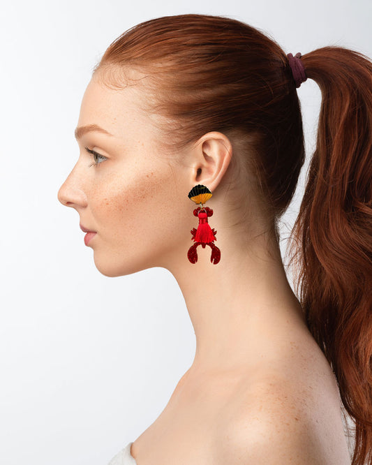 Dieser Ohrring ist gefertigt aus einem Edelstahl-Ohrsstecker in Form einer Venus-Muschel, einem rot glitzerndem Acryl-Lobster-Anhänger und einer roten Quaste.