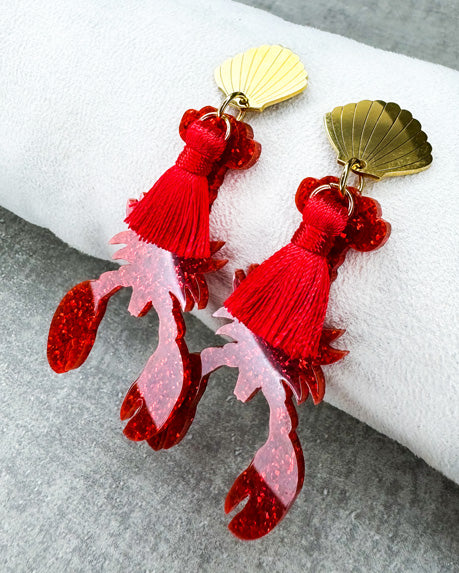 Dieser Ohrring ist gefertigt aus einem Edelstahl-Ohrsstecker in Form einer Venus-Muschel, einem rot glitzerndem Acryl-Lobster-Anhänger und einer roten Quaste.