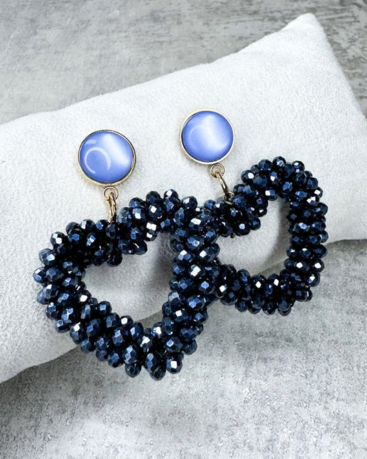 Dieser Ohrring ist gefertigt aus einem Cabochon-Stecker mit einem hellblau, glänzenden Stein und einem glitzernden Facetten-Herz-Anhänger in der Farbe "Silver Black.