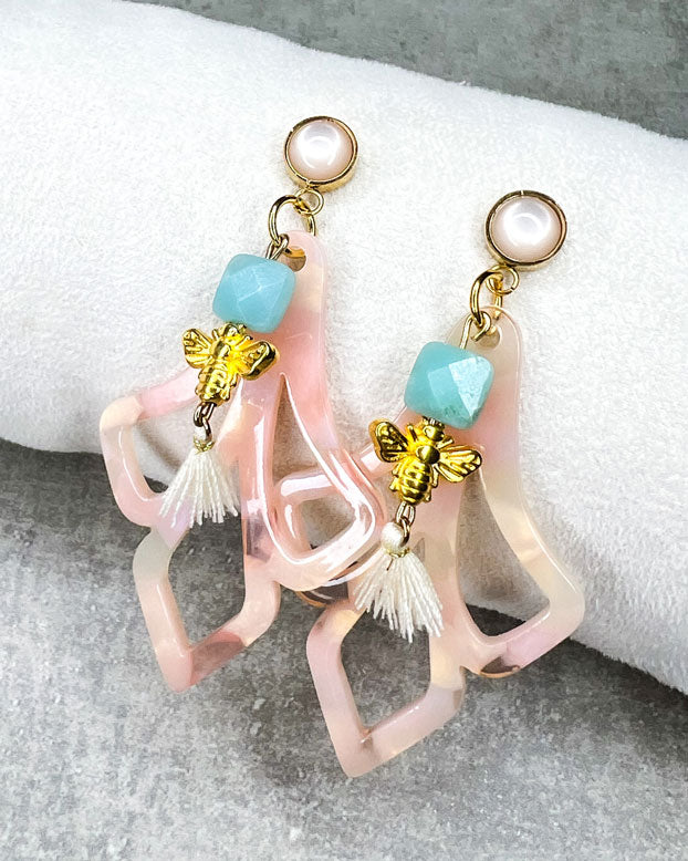 Diese Ohrringe sind gefertigt aus: einem goldfarbenem Cabochon Stecker mit einem baby-rosafarbenem Stein, einem Ornament-Anhänger in rosé-weiß, einer türkis-facettierten Jadeperle, einer gldfarbenen Bienenperle und einer cremefarbenen Quaste.