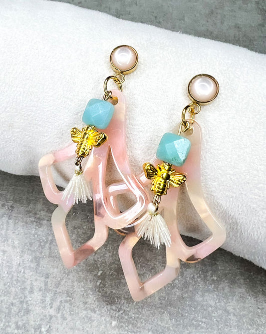 Diese Ohrringe sind gefertigt aus: einem goldfarbenem Cabochon Stecker mit einem baby-rosafarbenem Stein, einem Ornament-Anhänger in rosé-weiß, einer türkis-facettierten Jadeperle, einer gldfarbenen Bienenperle und einer cremefarbenen Quaste.