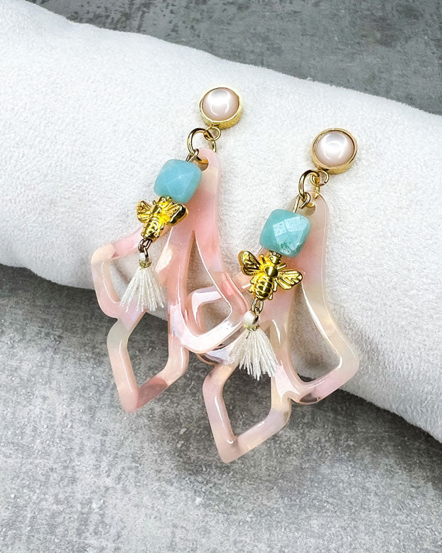 Diese Ohrringe sind gefertigt aus: einem goldfarbenem Cabochon Stecker mit einem baby-rosafarbenem Stein, einem Ornament-Anhänger in rosé-weiß, einer türkis-facettierten Jadeperle, einer gldfarbenen Bienenperle und einer cremefarbenen Quaste.