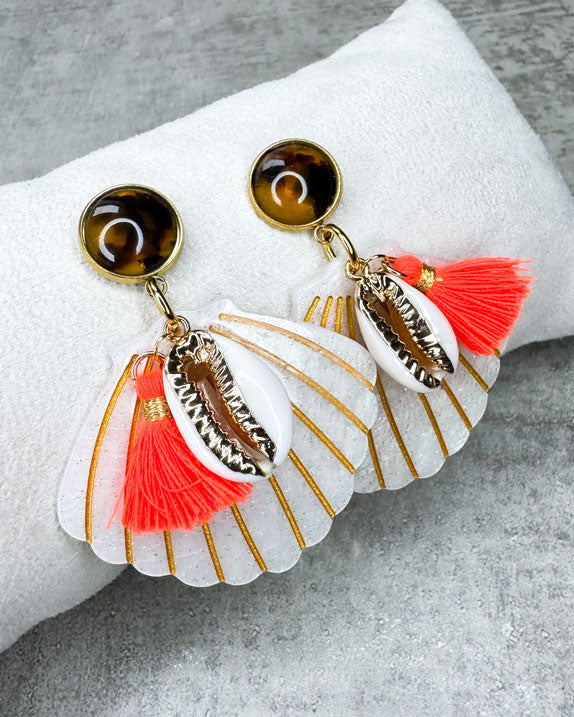 Dieser Ohrring ist gefertigt aus einem Polaris Steel Ohrstecker, einem Cabochon-Stein in Schildpatt-Optik, einem weiß glitzernden Muschel-Anhänger aus Acryl, einer gold verzierten Muschel als Anhänger und einer neon orangenen Quaste.
